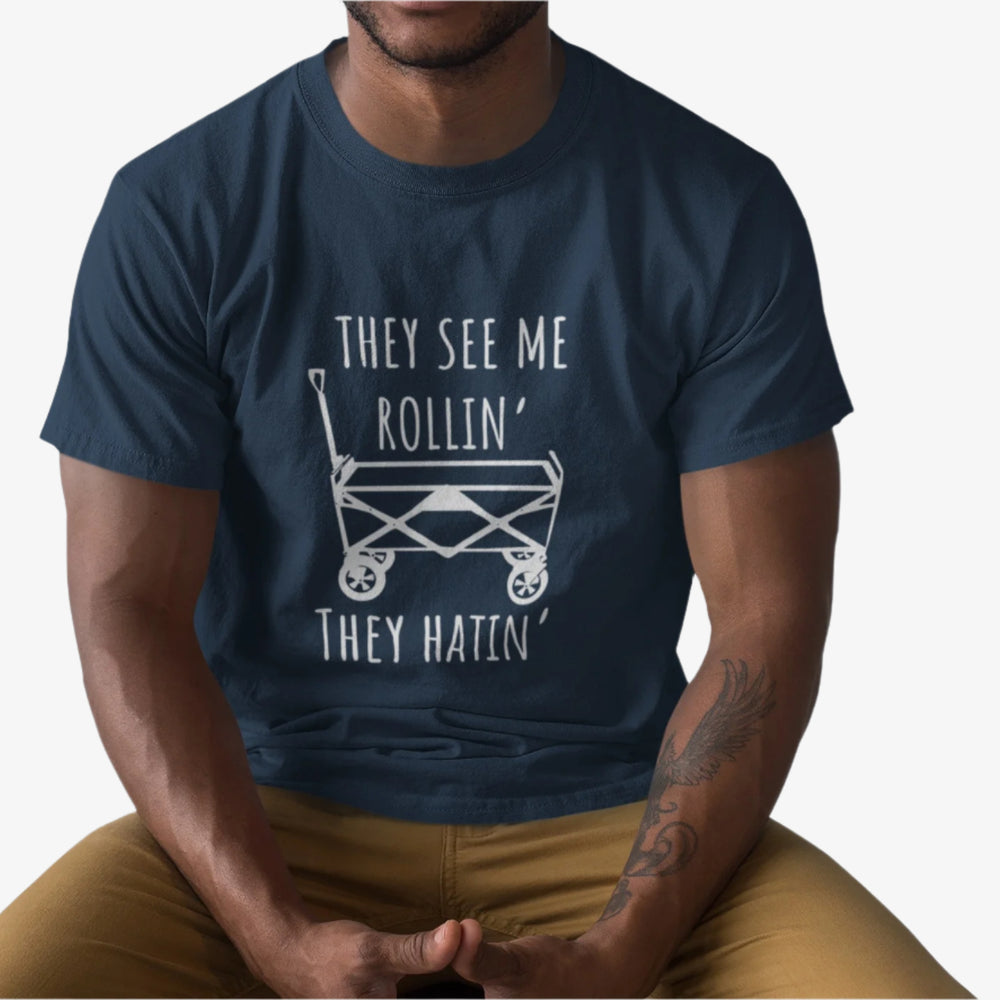 Person wearing a Navy blue t-shirt with white graphic playful design featuring a classic wagon graphic with the bold phrase, "They See Me Rollin' They Hatin'" on a white background